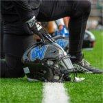 A football player dressed in black kneels on a turf field, resting a gloved hand on a black helmet with the blue �GV� logo for Grand Valley. Another helmet and players are visible in the background.